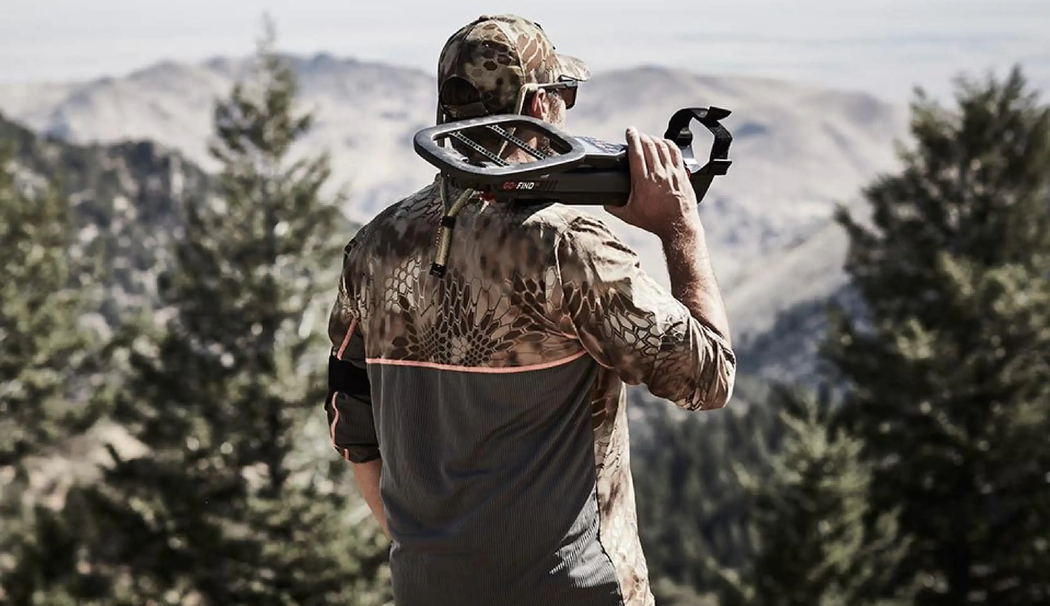 A man in a camouflage hunting shirt and cap stands outdoors in a mountain landscape, holding a folded black Minelab Go-Find 66 metal detector over his shoulder.