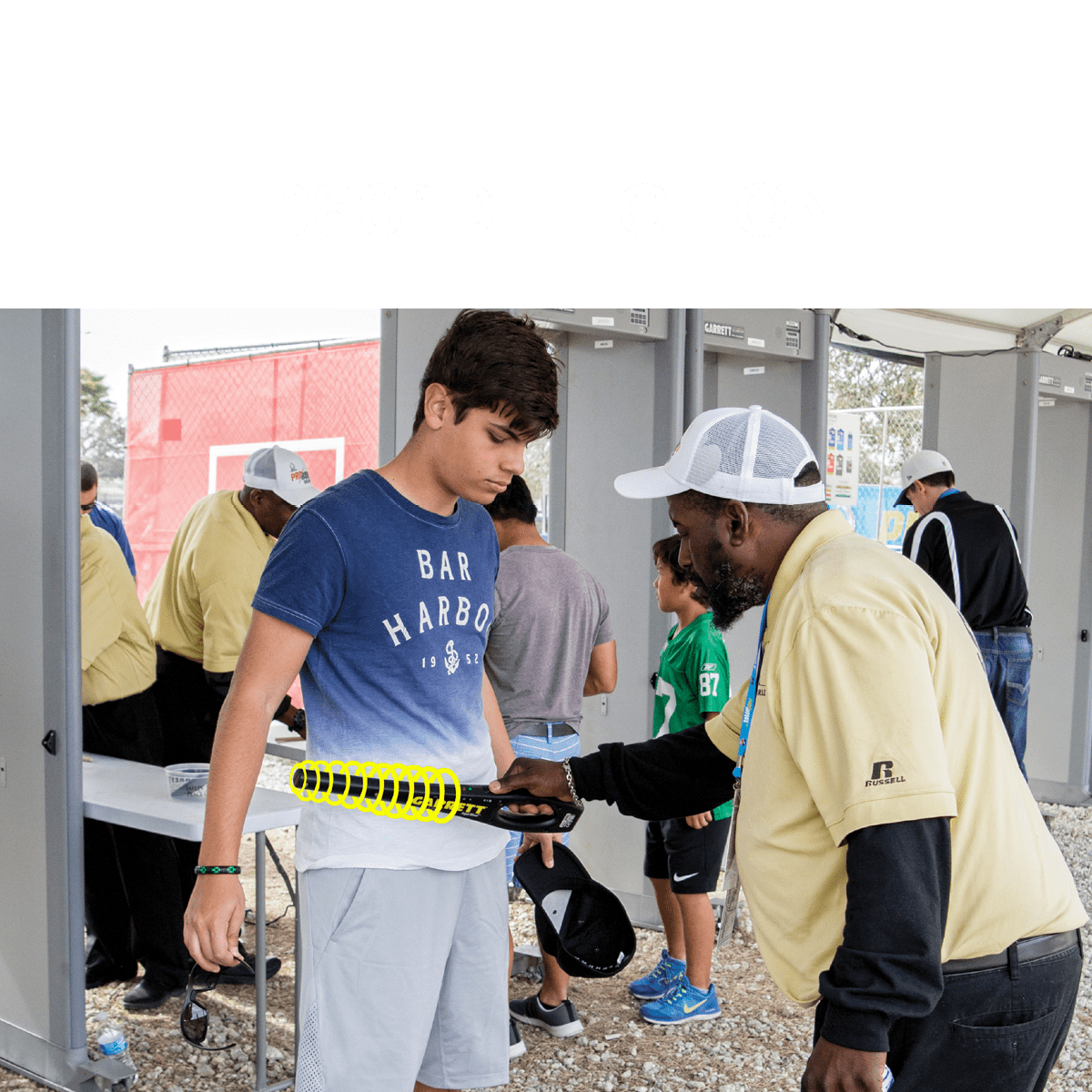 A man uses a yellow and black Garrett Superwand detector on a young man entering an outdoor event venue with other people visible in the background.